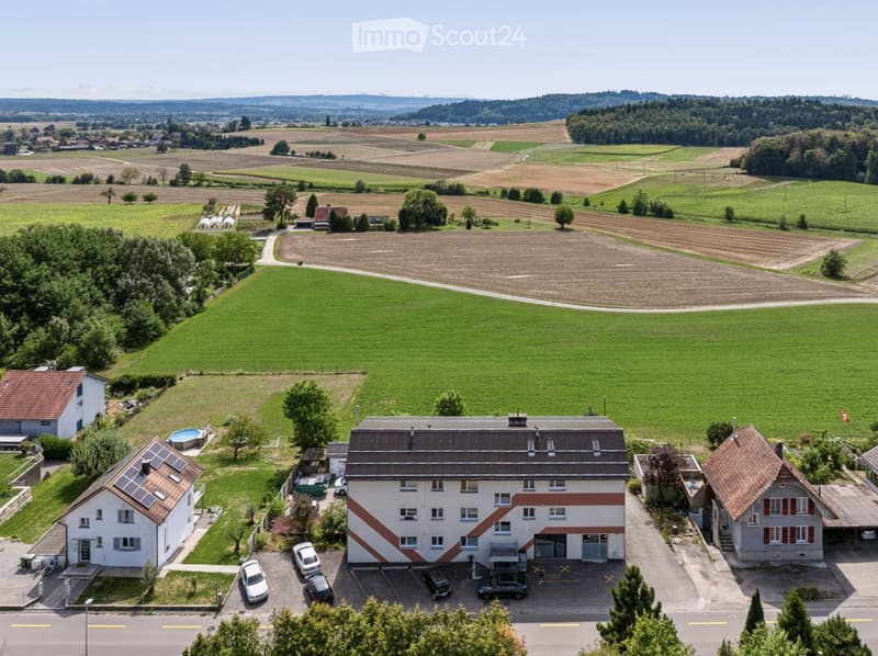 Aerial view of a residential area with multiple houses, a swimming pool, and surrounding green fields and trees.