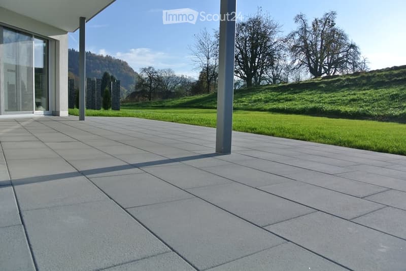 Modern patio with large concrete tiles, surrounded by green grass and trees, with a scenic mountain view in the background.