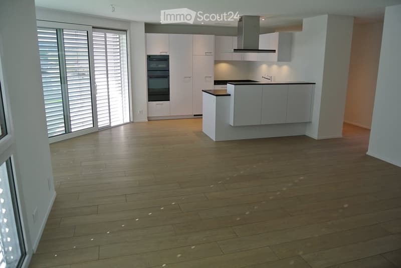 Empty kitchen with light wood flooring, white cabinetry, and built-in appliances including an oven and a range hood. Large windows with blinds provide natural light.
