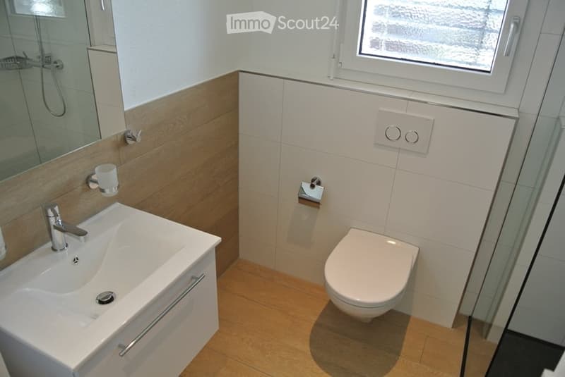 Modern bathroom with white tiles, wooden accents, a sink with a chrome faucet, a toilet, a mirror, and a window providing natural light.