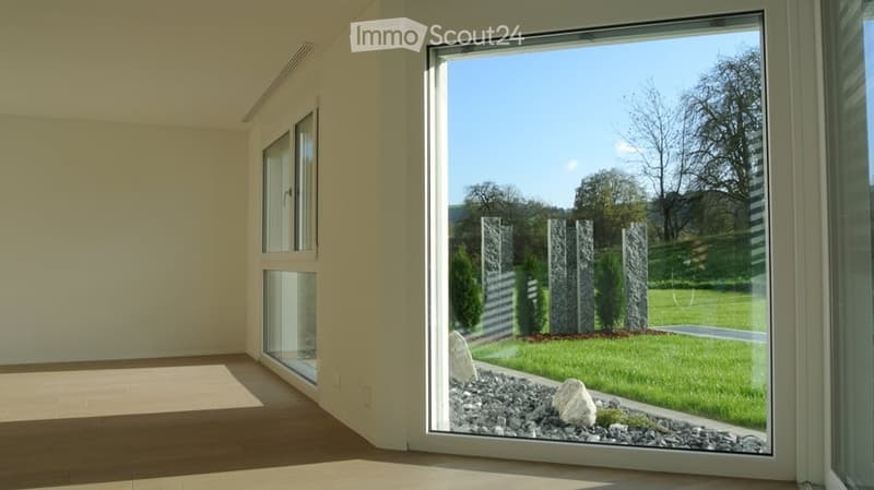 Bright, empty room with large windows and wooden flooring, offering a view of a landscaped garden area with rocks, shrubs, and trees.