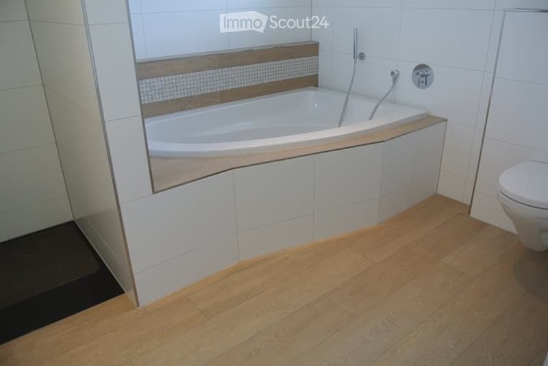 Modern bathroom with a white bathtub, wooden trim, white tiles, and a toilet. The floor is made of light wooden planks.