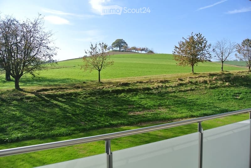Balcony overlooking a green, hilly landscape with scattered trees and a clear blue sky.