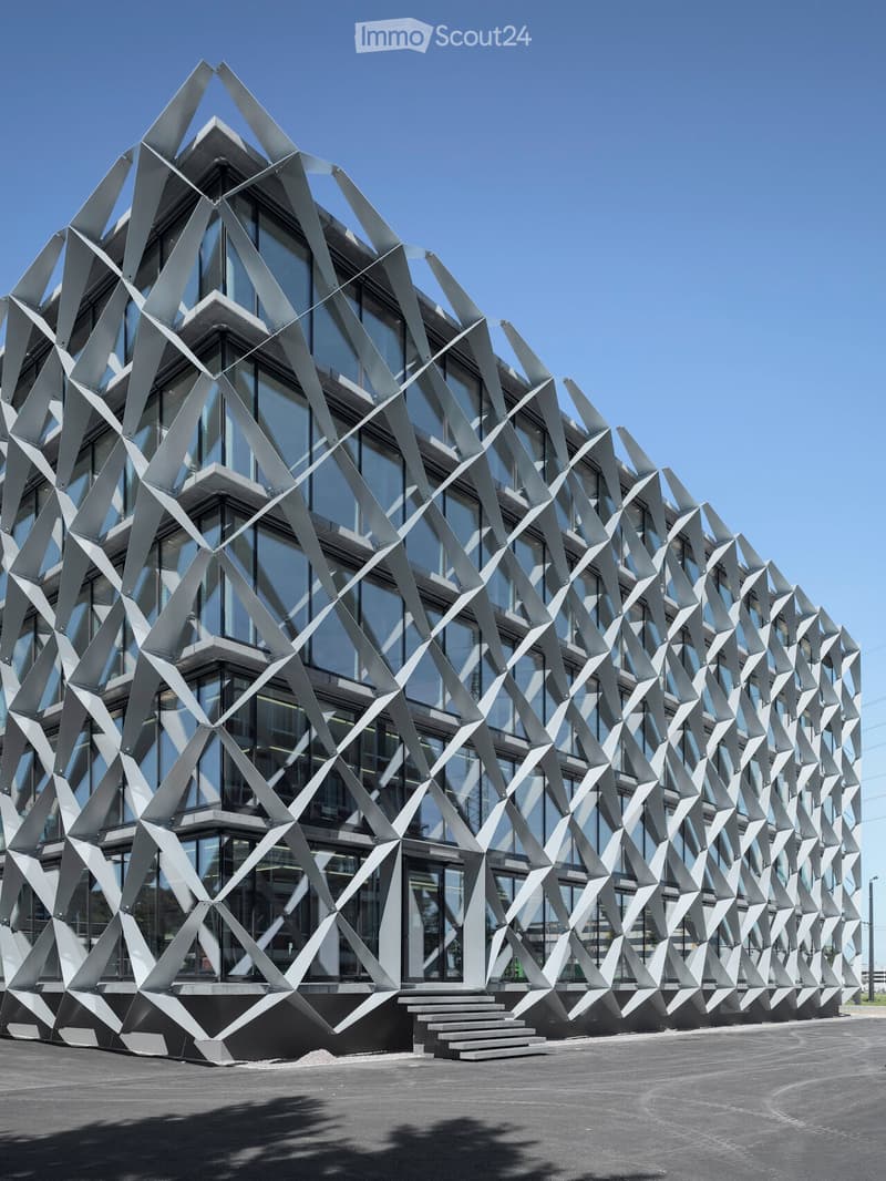 Modern building with a geometric, angular metal facade and large glass windows, featuring a raised ground floor and steps leading to the entrance.