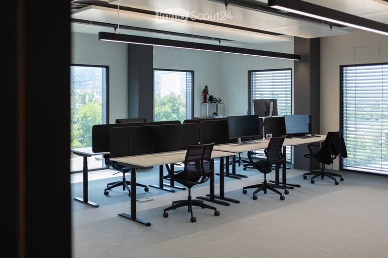Modern office space with multiple desks, ergonomic chairs, and computer screens arranged in rows. Large windows provide natural light, and the room features a clean, minimalist design with grey tones.