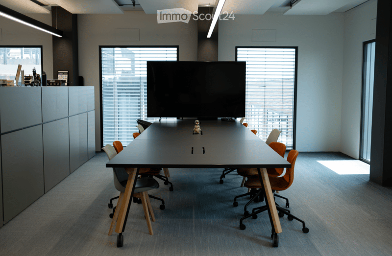 Modern conference room with a long rectangular table, multiple chairs, and a large screen at one end. The room has large windows with blinds and is well-lit with overhead lighting.