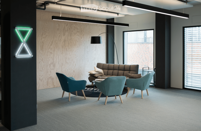 Modern, well-lit lounge area with a grey sofa, teal armchairs, a coffee table, and a floor lamp. The space features large windows and a sleek, contemporary design.