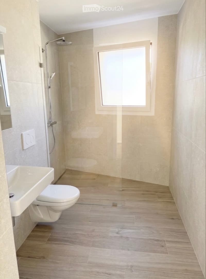 Modern bathroom with light-colored tiles, wooden flooring, a toilet, sink, and a glass-enclosed shower with a handheld showerhead.