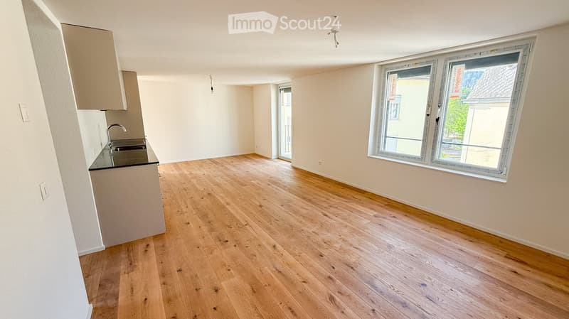Empty room with light wooden flooring, white walls, and a small kitchen area with a sink and cabinets. Large windows and a door leading to a balcony or outdoor area are visible.