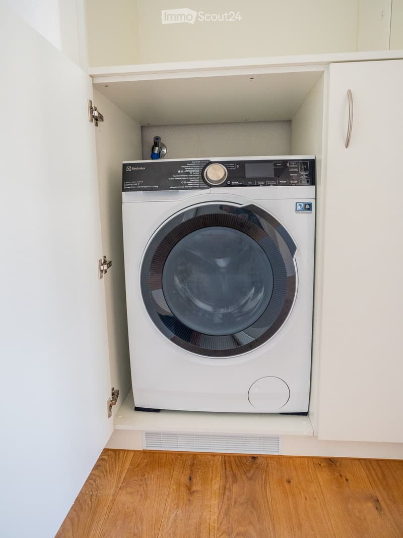 A white washing machine is installed in a built-in cabinet with wooden flooring.