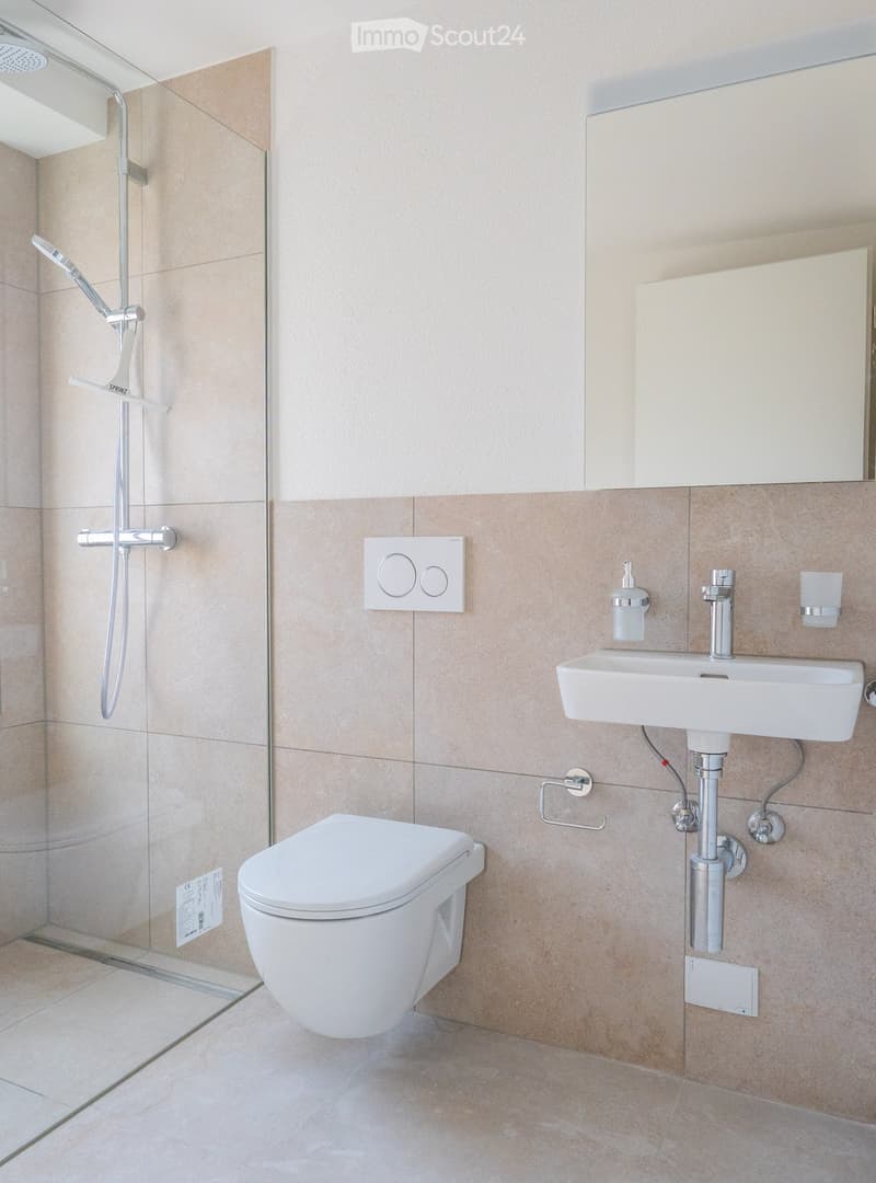 Modern bathroom with beige tiles, a toilet, a sink with a faucet, a shower with a handheld sprayer, a mirror, and a towel rack.