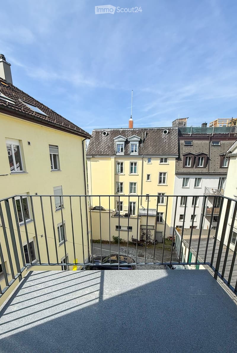 Balcony overlooking a cityscape with multiple residential buildings, metal railing, and parked cars below.