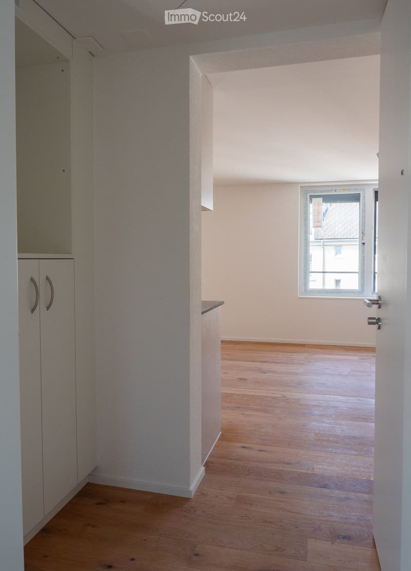 Empty room with wooden flooring, white walls, and a window with a view of a building outside. The room features built-in cabinets with handles on the left side.