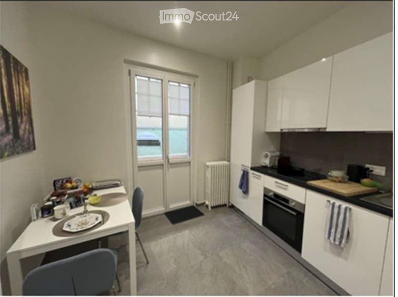 Modern kitchen with white cabinets, black countertops, and stainless steel appliances. Features a dining table with chairs, a radiator, and a window with a view of the outside.