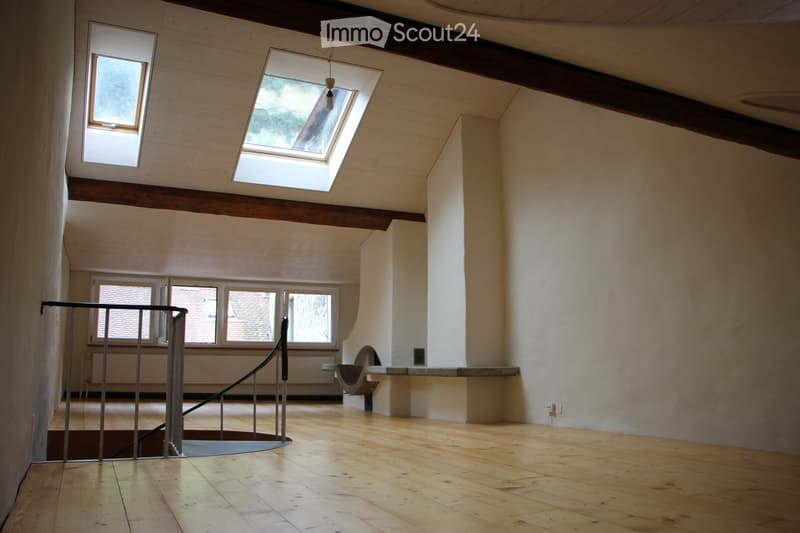 Empty room with wooden flooring, exposed wooden beams on the ceiling, and large windows including a skylight. A metal staircase railing is visible leading to another level.