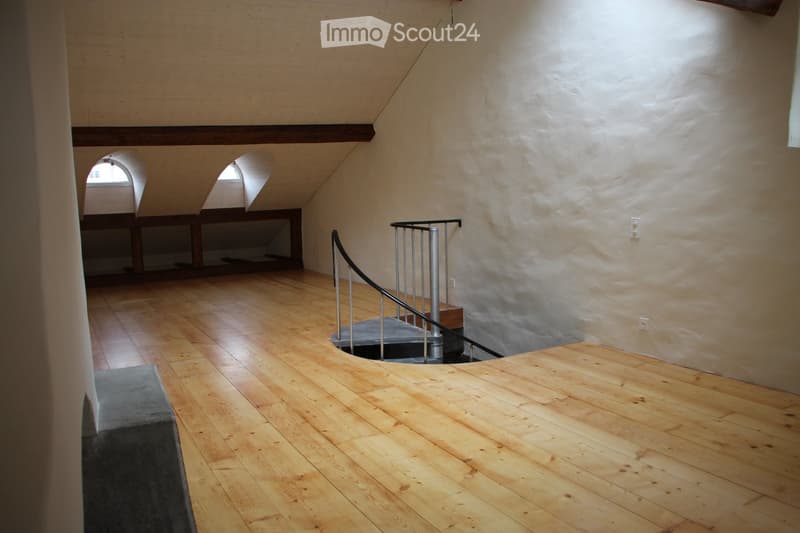 Empty room with wooden flooring, exposed wooden beams on the ceiling, and a modern metal staircase leading upwards.