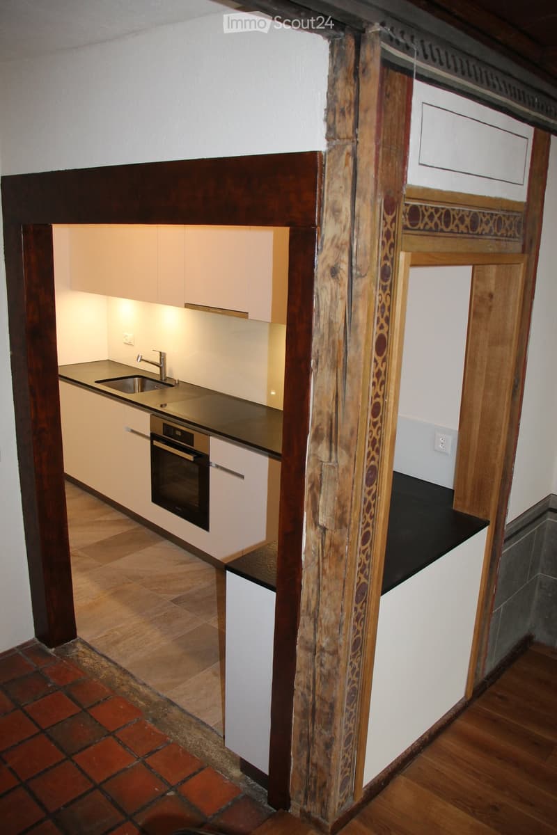 Modern kitchen with white cabinets, stainless steel sink, and oven. The space features a wooden pillar and a mix of tiled and wooden flooring.