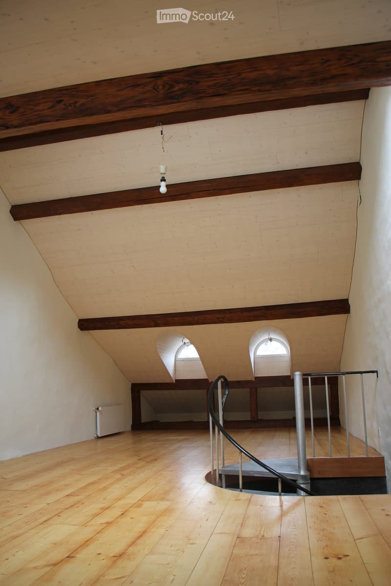 Empty wooden hallway with exposed wooden beams, a spiral staircase leading downwards, and arched windows providing natural light.
