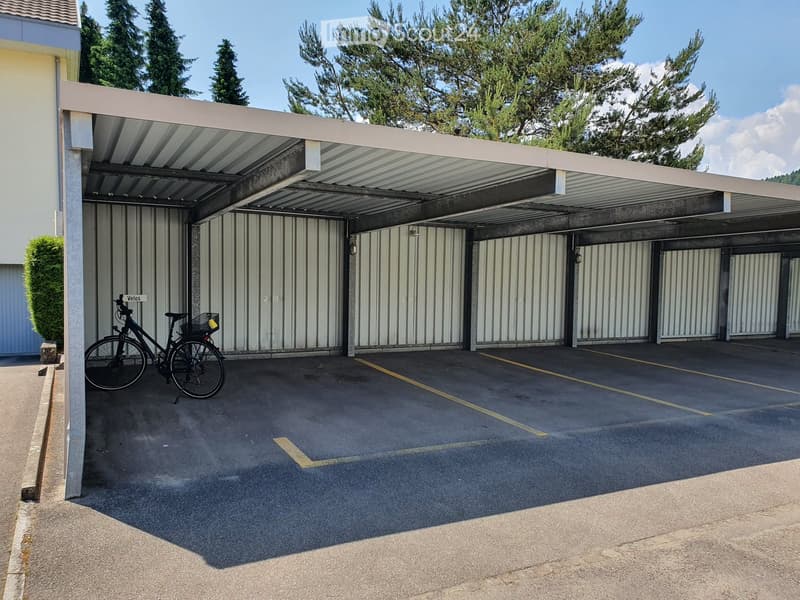Covered parking area with concrete floor, metal roof, and several closed garage doors. A bicycle is parked on the left side.