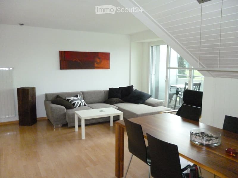 Modern living and dining area with a grey sectional sofa, coffee table, dining table with chairs, and a painting on the wall. The space features wooden flooring and large windows with blinds.