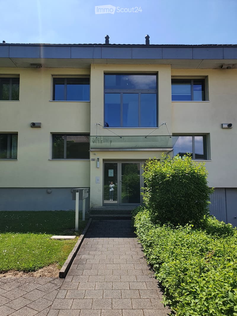 Two-story building with light green facade, large windows, and a central entrance. The entrance features a glass door and a small pathway leading to it, flanked by neatly trimmed bushes and a small lawn area.