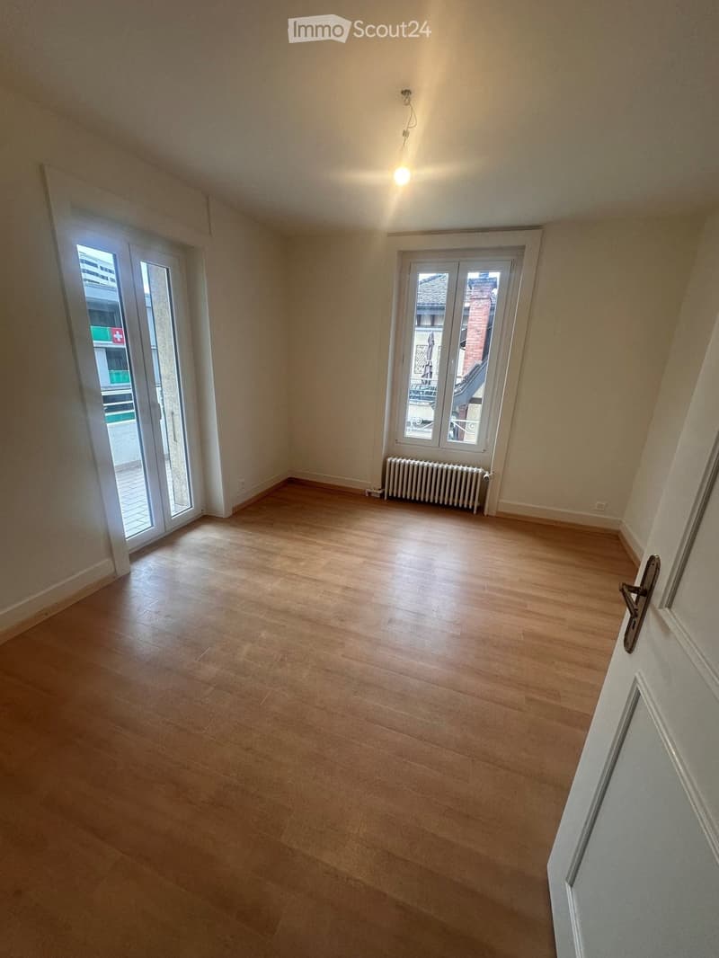 Empty room with wooden flooring, white walls, and large windows with sliding doors leading to a balcony.