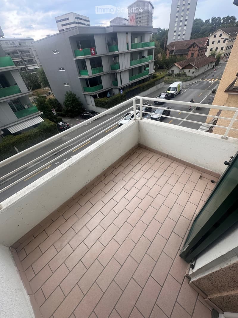Balcony with tiled floor, white railing, overlooking a street with parked cars and buildings.