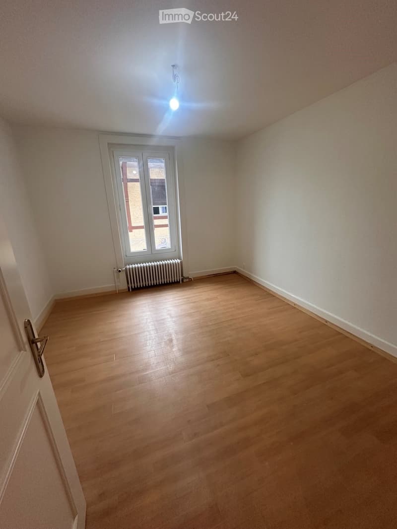Empty room with wooden flooring, white walls, a window with a radiator below, and a door on the left side.
