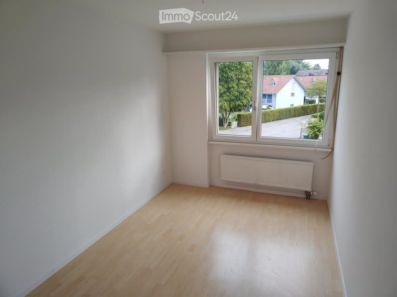 Empty room with light wooden flooring, white walls, and a large window offering a view of the street and neighboring houses.