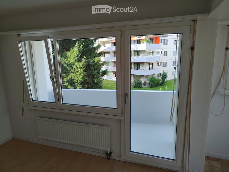 Empty room with wooden flooring, white walls, large windows, radiator, and a view of a green area and neighboring buildings.