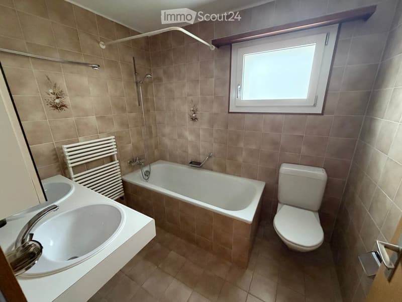 Bathroom with beige tiles, a bathtub, toilet, sink, and a radiator. A window provides natural light.