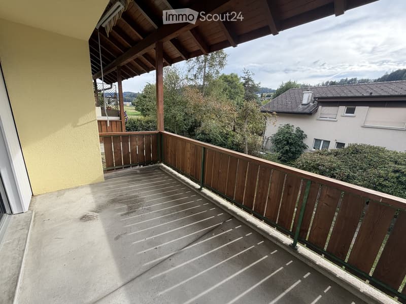 Balcony with wooden railing, concrete floor, and a view of a residential area with trees and houses.