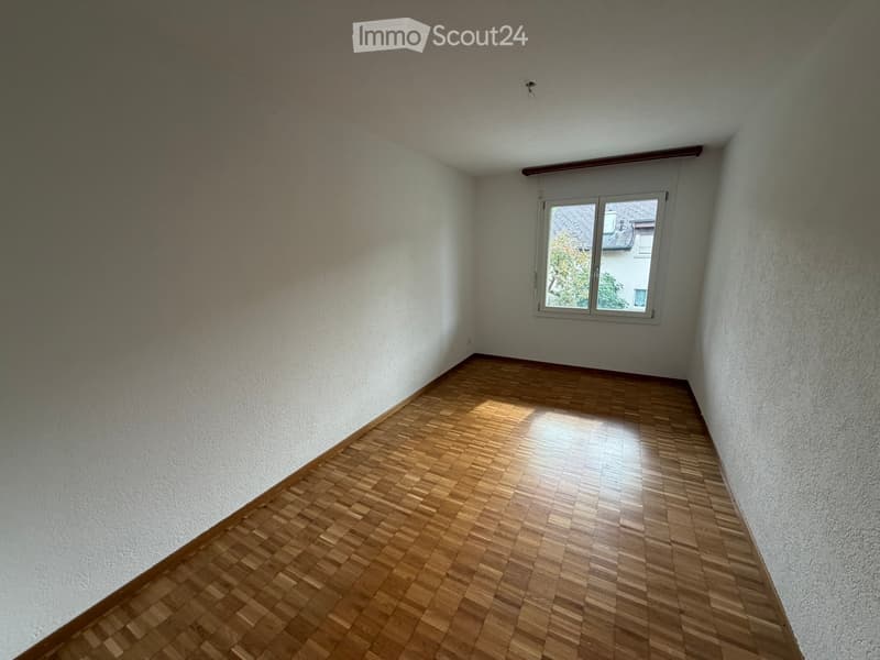 Empty room with wooden flooring and white walls, featuring a single window with a view of a neighboring building.