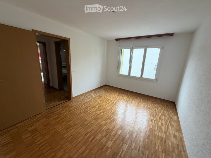 Empty room with wooden flooring, white walls, and a large window with white frames. The room has an open doorway leading to another area.