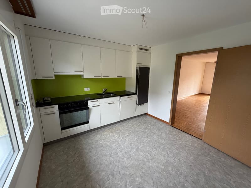 Modern kitchen with white cabinets, green backsplash, and black countertops. Features an oven, refrigerator, sink, and faucet. Open doorway leads to a wooden-floored room.