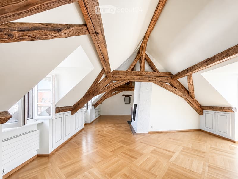 An attic space with exposed wooden beams, wooden herringbone flooring, white walls, and built-in white cabinets along the walls. The room is empty, with no furniture or personal items visible.