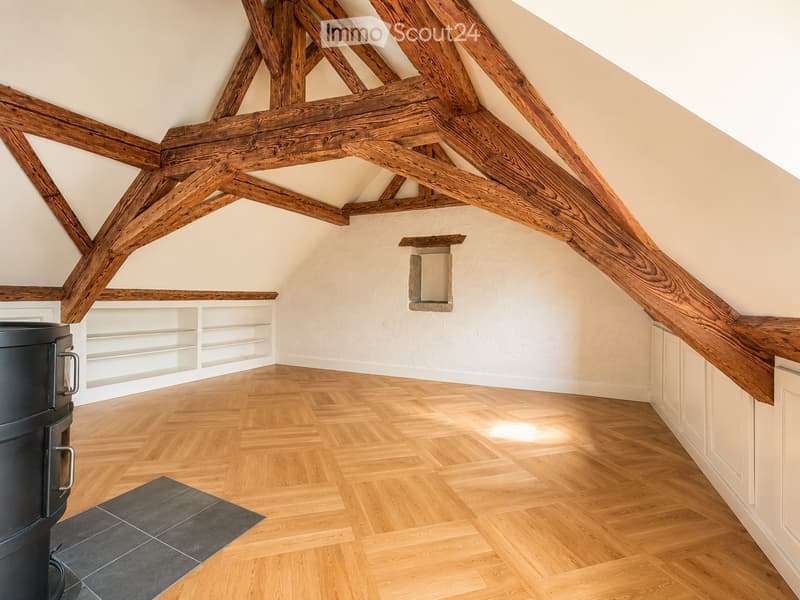 An empty attic space with exposed wooden beams, wooden flooring, built-in white shelving, and a black wood-burning stove on a tiled platform.