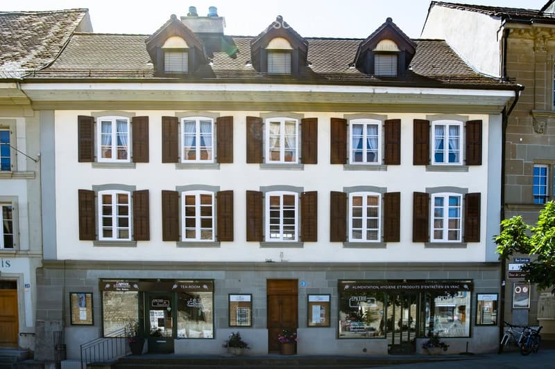 Three-story building with white facade, multiple windows with brown shutters, and commercial storefronts on the ground floor including a tea room and grocery store.