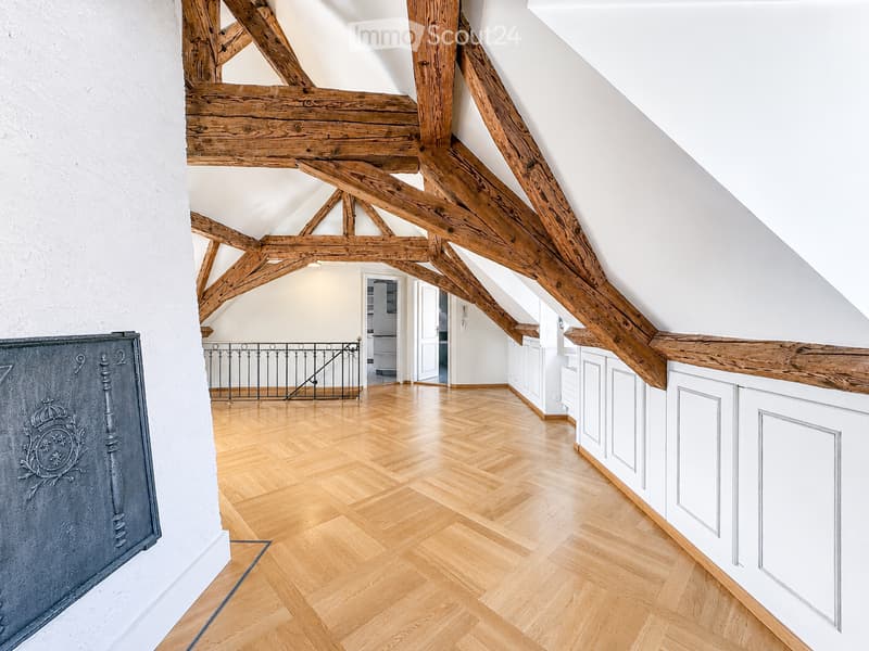 Bright, airy hallway with exposed wooden beams and herringbone wooden flooring, leading to an open area with white cabinetry and a metal railing.