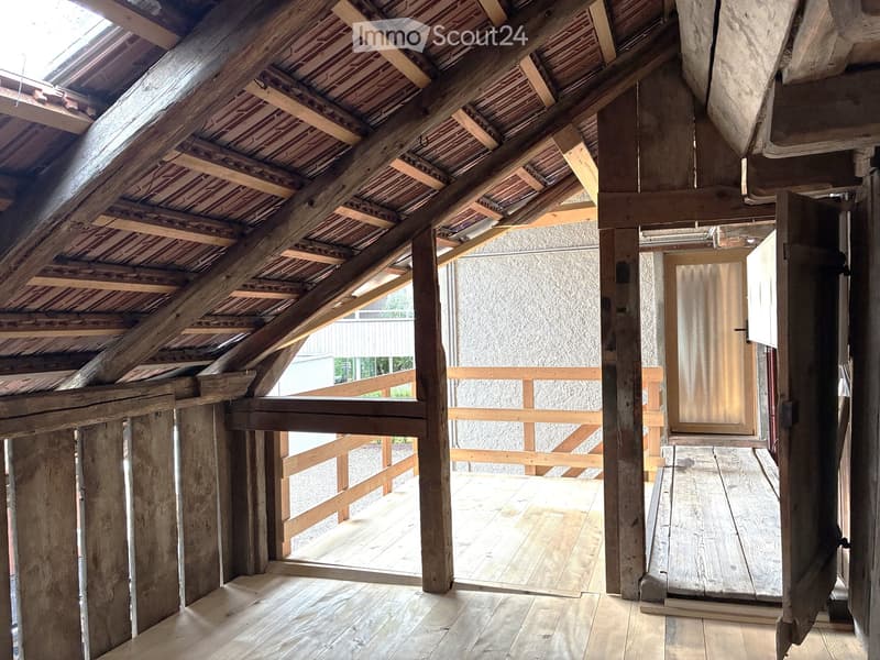 An attic space with exposed wooden beams and rafters, wooden floor, and a doorway leading to a wooden deck area.
