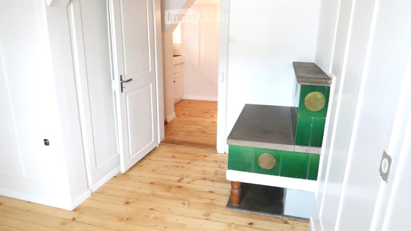 A narrow hallway with wooden flooring, white walls, and a small green and grey staircase with gold accents leading upwards.