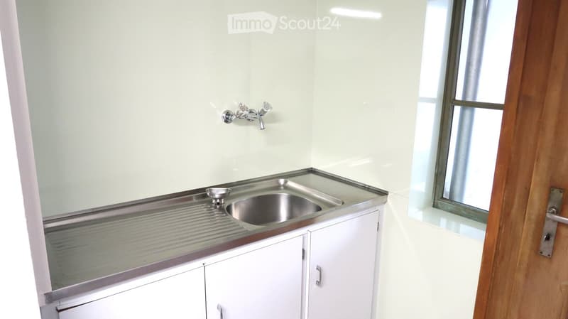 Empty room with white walls, a stainless steel sink, and a faucet. The room has a wooden door and a window with a metal frame.