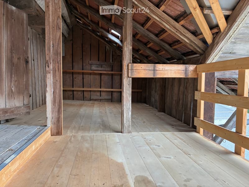 An empty wooden attic space with exposed wooden beams and a slanted wooden ceiling. The floor is made of light-colored wooden planks, and the walls are lined with wooden planks and beams.