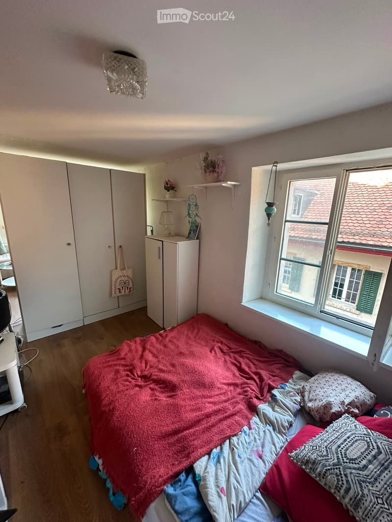 A cozy bedroom with a double bed covered in a red blanket and pillows, a wardrobe, a small table with a lamp, and a window overlooking a tiled roof.