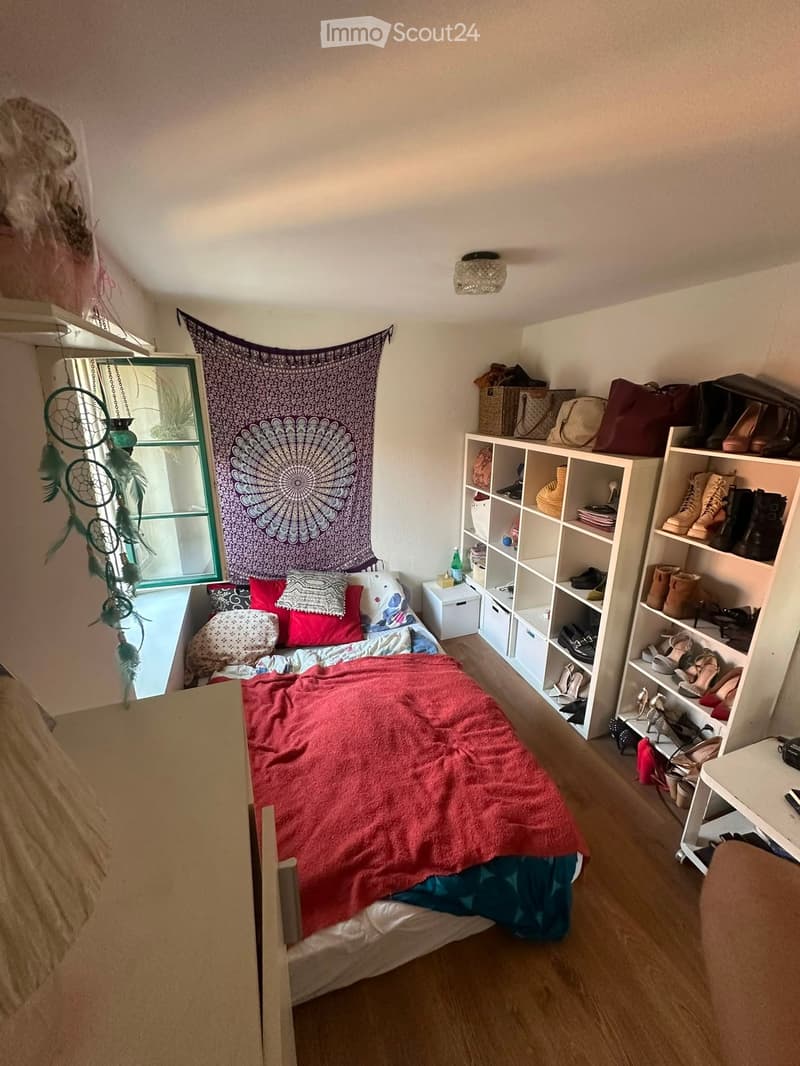 Cozy bedroom with a single bed, colorful pillows, red blanket, shelves filled with shoes and storage boxes, and a dreamcatcher near the window.