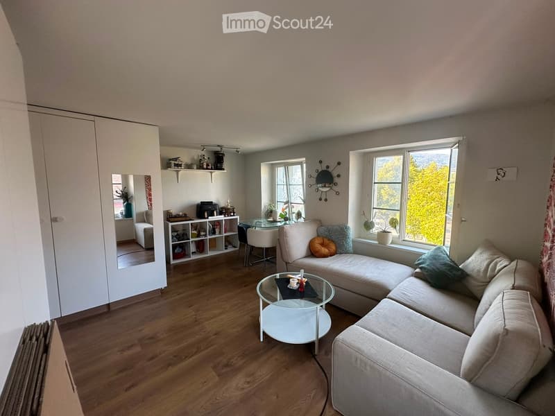 Modern living room with light-colored wooden flooring, a large sectional sofa, a glass coffee table, a small dining area with a glass table and chairs, and a shelf with decorative items.