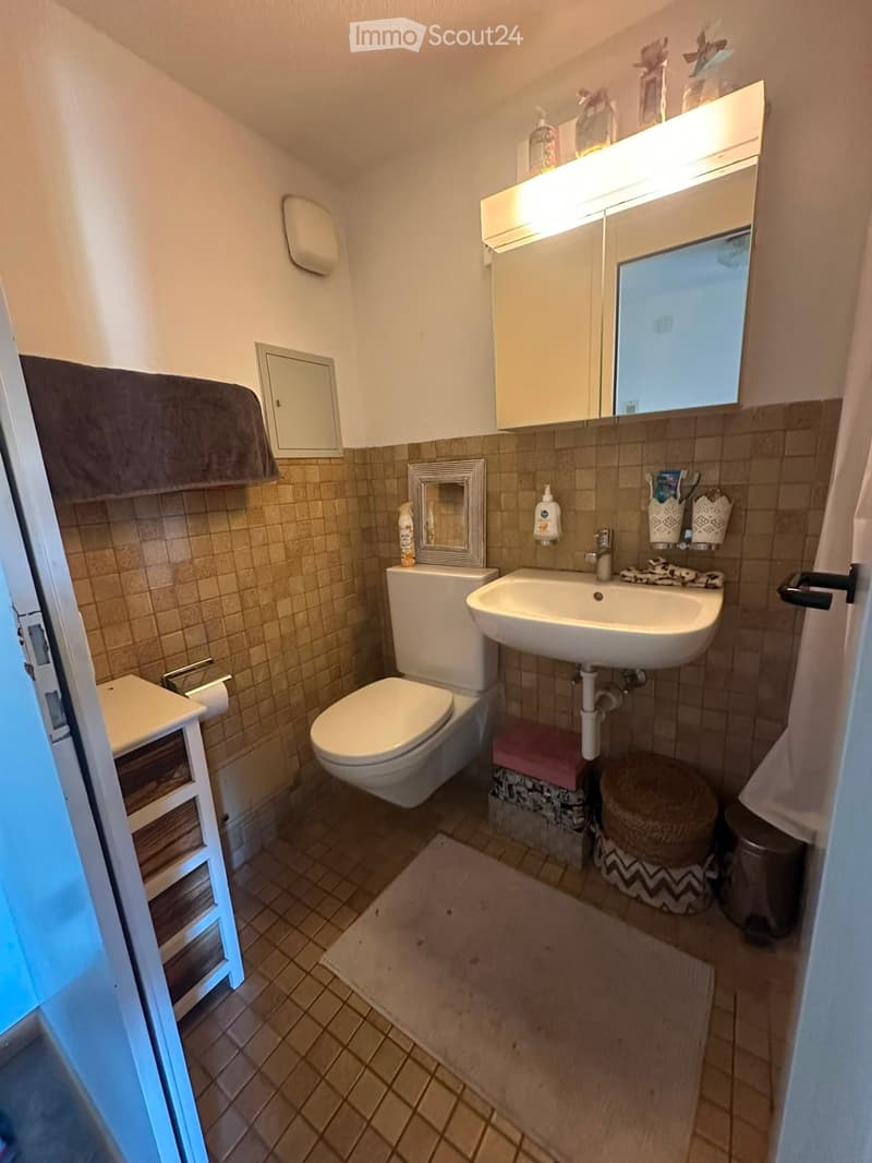 Compact bathroom with beige tiles, a toilet, a sink with a mirror cabinet, a small wooden cabinet, and a towel on a rack.
