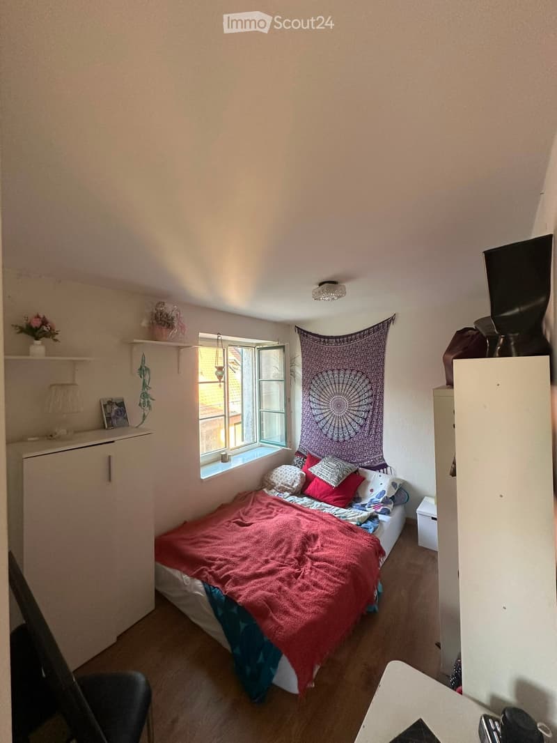 Cozy bedroom with a single bed, colorful pillows, red blanket, wooden floor, white cabinets, a small table, and a wall tapestry. A window with a view of a rooftop is visible.