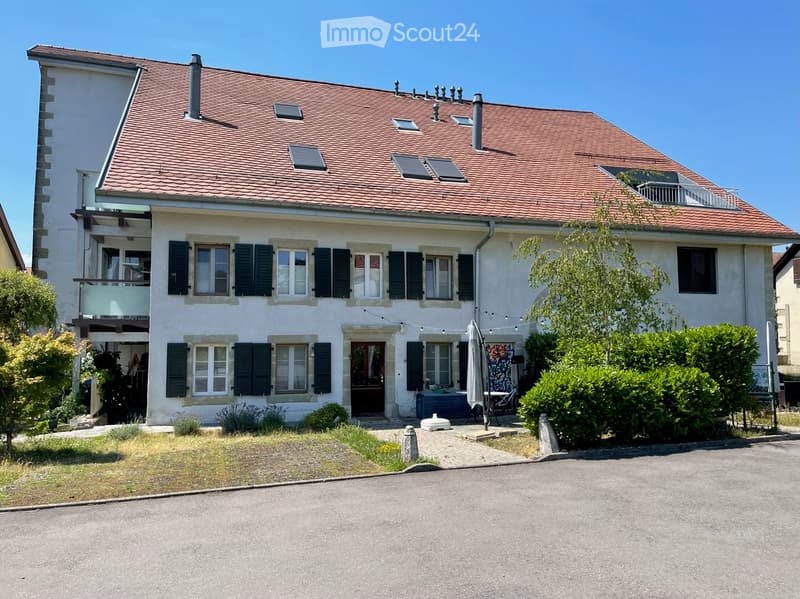 Two-story house with red-tiled roof, white walls, multiple windows with shutters, and a balcony on the upper level. The front yard includes a small patio area with outdoor furniture.