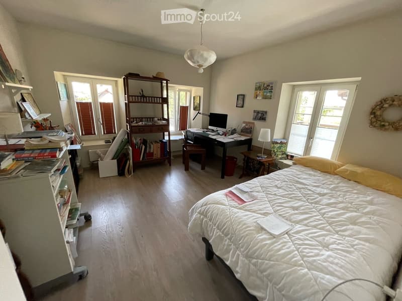 Bright bedroom with wooden flooring, large windows, a bed with white bedding, a desk with a computer, a bookshelf, and several picture frames on the walls.